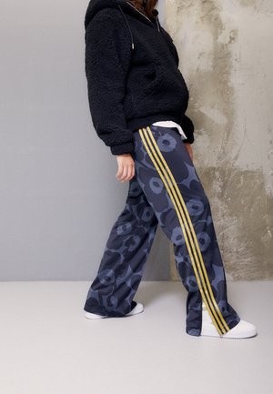 Personne portant un sweat à capuche en polaire noire et un pantalon bleu marine avec un grand motif floral et des rayures jaunes marchant à l'intérieur sur un sol clair.