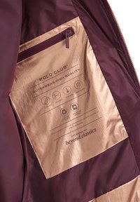 Maroonfärgad jackas insida med en metallicguldig ficka som visar Polo Club-varumärket och ikoner för ultralätt, skräddarsydd passform, extra varm, vattenresistent, återvinningsbar.