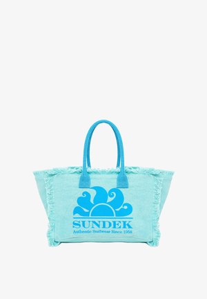 Borsa a tracolla azzurro chiaro con bordo frangiato, che presenta una grande grafica di un sole blu e la scritta "SUNDEK Authentic Surfwear Since 1968".