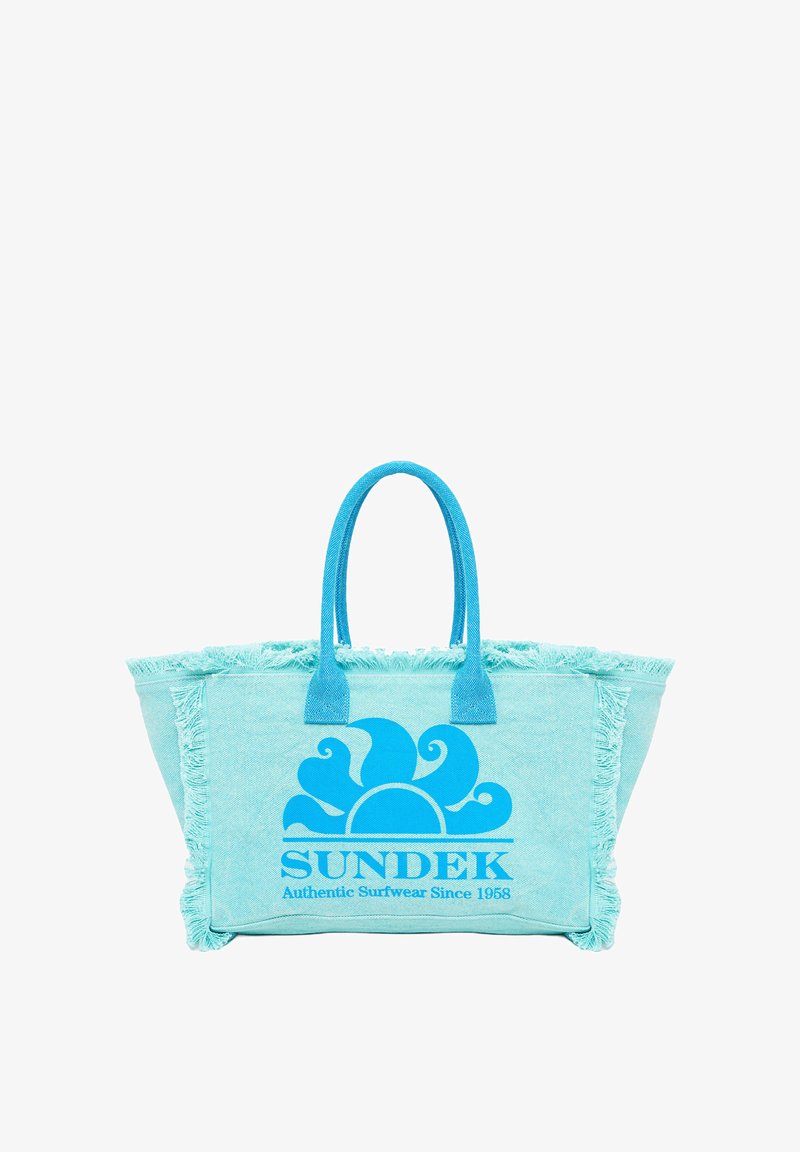 Bolso tote azul claro con un borde de flecos, que presenta un gran gráfico de un sol azul y el texto "SUNDEK Ropa de Surf Auténtica Desde 1968".