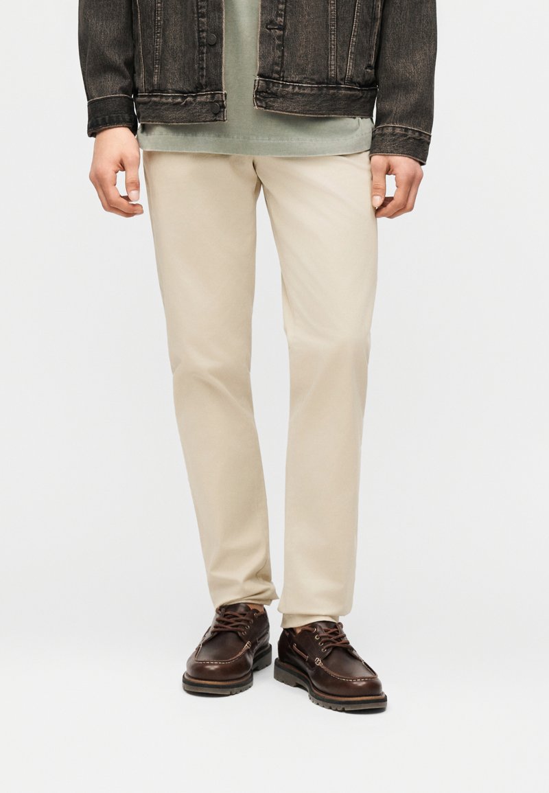 Homme portant un pantalon beige, des chaussures en cuir brun foncé, une chemise verte et une veste en jean noire, se tenant devant un fond blanc.