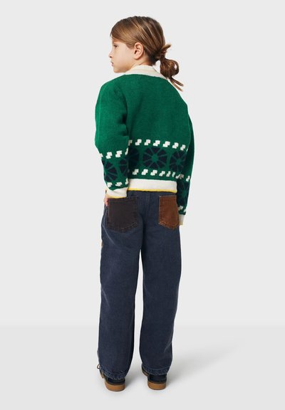 Grüner Strickpullover mit weißem und marineblauem Muster, kombiniert mit dunkelblauen Jeans, die braune Lederapplikationen haben.