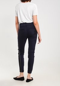 En person med lockigt hår bär en ljusgrå kortärmad topp och marinblå byxor med sidsömmar, sedd bakifrån, stående på en ljus bakgrund.