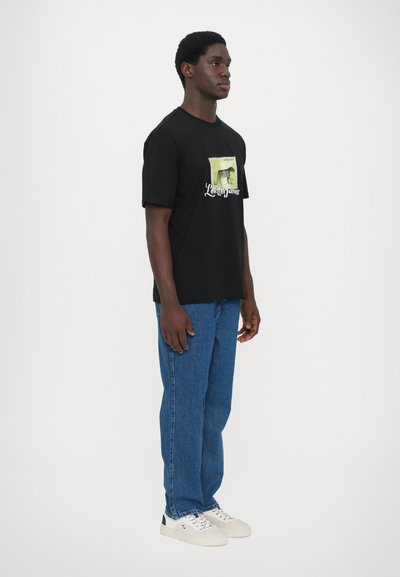 T-shirt en coton noir avec un imprimé graphique sur le devant, associé à un jean en denim bleu et des baskets blanches avec des accents noirs.