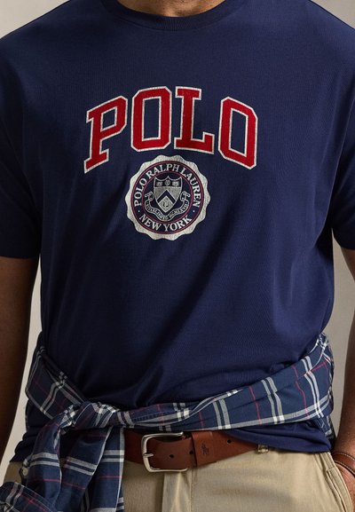 Μπλε σκούρο Polo Ralph Lauren T-shirt με κόκκινο λογότυπο και έμβλημα, καρό πουκάμισο δεμένο γύρω από τη μέση, μπεζ παντελόνι και καφέ δερμάτινη ζώνη.