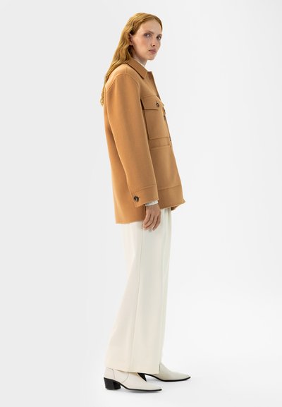 Helle braune Wollmischung Jacke mit zwei Brusttaschen, Knopfverschluss und lockerer Passform, kombiniert mit weißen weiten Hosen und spitzen Stiefeln.