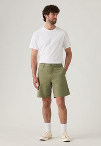 Shorts en coton vert à coupe droite, poches latérales, associés à un t-shirt blanc uni et des chaussures beiges, debout sur un fond neutre.
