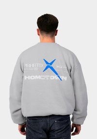 Pelēka džemperis ar lielu zilu grafiku un dažādiem tekstiem mugurpusē. Teksts ietver "PRHBTD" un "HOMETOWN" baltā krāsā. Garās piedurknes.