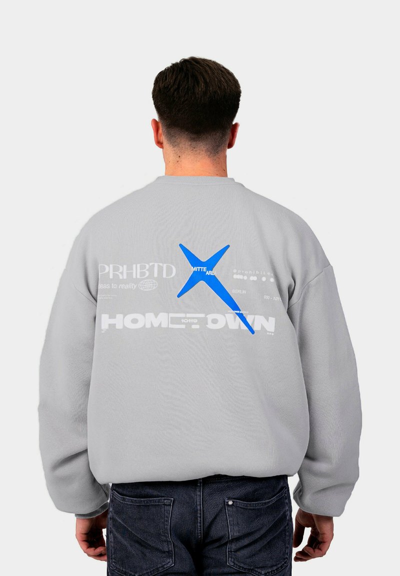 Pelēka džemperis ar lielu zilu grafiku un dažādiem tekstiem mugurpusē. Teksts ietver "PRHBTD" un "HOMETOWN" baltā krāsā. Garās piedurknes.