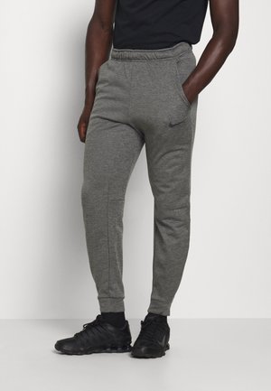 Homme portant un pantalon de survêtement Nike gris avec taille élastique et poches, chemise noire, chaussettes noires et chaussures de sport noires, debout contre un mur blanc.