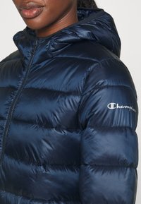 Kvinna som bär en marinblå puffjacka med huva, med "Champion"-logotypen på den övre ärmen, dragkedjan delvis synlig, läppar och haka syns.