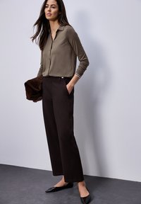 Femme debout portant un chemisier marron, un pantalon large sombre, des ballerines noires, tenant une veste marron pliée, devant un mur clair uni.