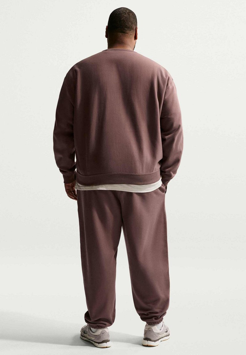 Man met de rug naar de camera, draagt een losse mauvekleurige trui en bijpassende broek over een wit shirt, met grijze sneakers op een effen achtergrond.