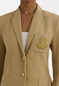 Blazer beige en tissu texturé, à deux boutons, avec une poche poitrine ornée d'un logo brodé en or. Poches verticales de chaque côté.