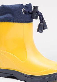 Geel rubberen regenlaars met een marineblauwe stoffen rand en zwarte hardware. Glad oppervlak met een gebogen vorm en een duurzame zool voor grip.