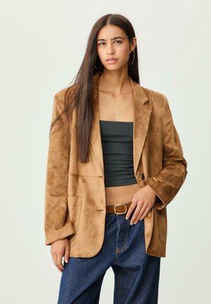 Femme aux longs cheveux bruns portant de grandes boucles d'oreilles en or, un blazer en suède marron, un top sans manches noir, un jean bleu et une ceinture marron.