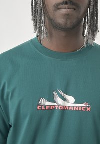 Green cotton t-shirt featuring a graphic of a flying bird and the text "CLEPTOMANICX" in pink, round neckline, and short sleeves.