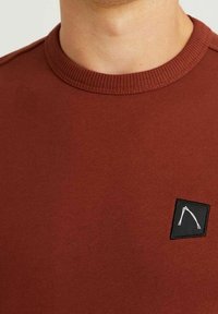 Felpa bordeaux con colletto a coste e una toppa quadrata nera con un design bianco sul davanti. Tessuto morbido e texturizzato.