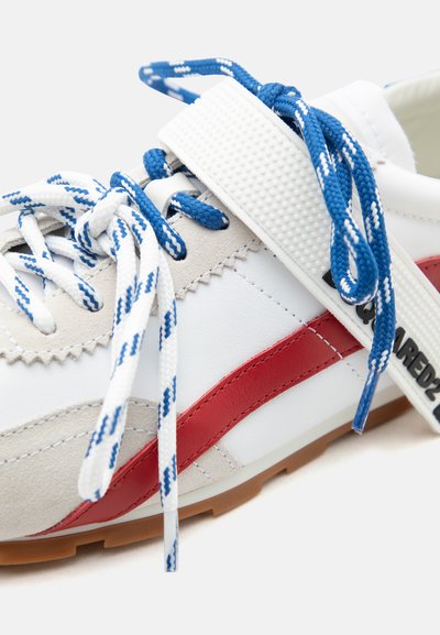 Baskets blanches et beiges avec une bande rouge, des lacets bleus et blancs à motifs, et une étiquette blanche portant la mention "DSQUARED2" attachée à un lacet.