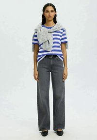 T-shirt azul e branco às riscas, suéter cinza claro drapeado sobre os ombros, calças largas cinza de cintura elevada, sapatos pretos, pose de pé.