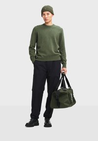 Maglione in lana verde oliva, pantaloni cargo neri, stivali neri. Tiene una borsa da viaggio verde scuro con cinghie e fibbie nere, con dettagli bianchi.