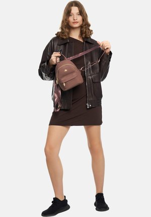 Kvinde iført en mørkebrun læderjakke og kjole, der holder en lille lilla rygsæk med guldlynlås og biemblem, stående mod en hvid baggrund.