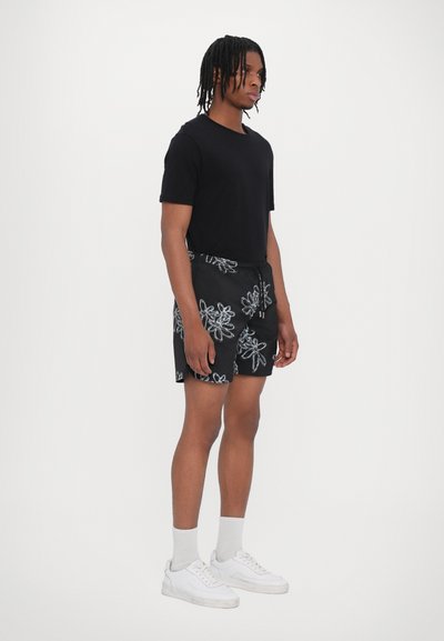 Schwarzes T-Shirt kombiniert mit schwarzen Shorts mit weißen Blumenmustern, getragen mit weißen Sneakern und hellgrauen Socken.