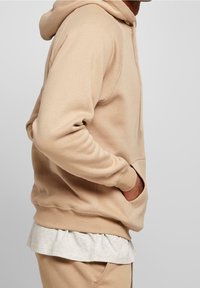 Beige hoodie met een kangoeroezak, ribgebreide manchetten en zoom. Draagt over een lichtgrijs T-shirt, met een relaxte pasvorm en zachte textuur.