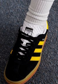 Zapatilla Adidas negra y amarilla con tres rayas, hecha de materiales de ante y malla, suela de goma y cordones negros. Media blanca parcialmente visible.