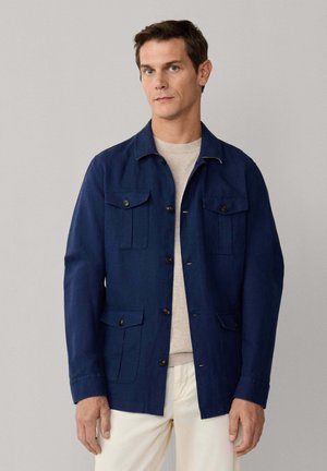 Man draagt een donkerblauwe jas met knopen en vier zakken met kleppen, over een beige overhemd en crèmekleurige broek, staand tegen een effen grijze achtergrond.