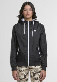 Black windbreaker jacket with a zippered front, white accents, and a small butterfly logo on the chest. Features a hood and elastic cuffs.