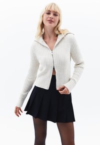 Kremowy sweter w prążki z zapięciem na zamek i kapturem, zestawiony z czarną spódnicą plisowaną. Sweter ma długie rękawy i dopasowany krój.