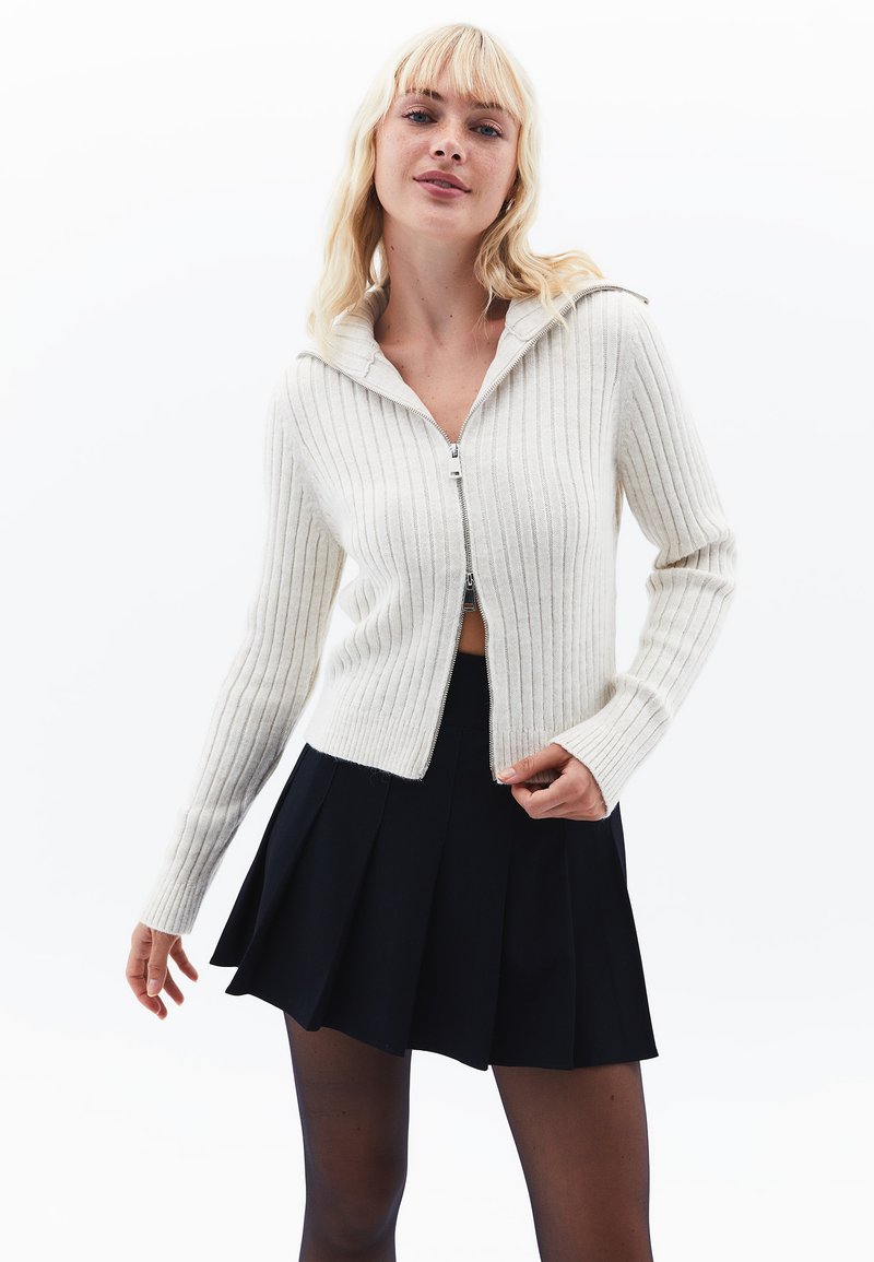 Kremowy sweter w prążki z zapięciem na zamek i kapturem, zestawiony z czarną spódnicą plisowaną. Sweter ma długie rękawy i dopasowany krój.