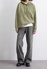 Olivengrön hoodie med känguruficka och dragsko, lagd över en vit skjorta; ihop med löst sittande, urtvättade grå jeans och svarta skor.