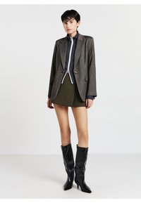 Blazer grigio con texture e bottoni dorati, indossato sopra un top blu navy con zip e una minigonna oliva, abbinato a stivali neri lucidi fino al ginocchio.