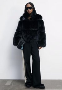 Veste en fausse fourrure noire avec de larges manches et un col haut, assortie à un pantalon noir avec une bande latérale blanche et des chaussures noires épaisses.