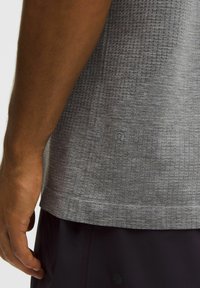 Tricou sport gri cu material texturat și model subtil. Prezintă un logo pe partea stângă jos. Mâneci scurte și tiv drept.