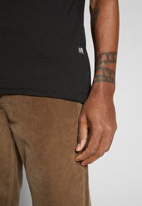Black cotton t-shirt with a small logo tag on the hem, paired with textured brown corduroy pants, showcasing vertical ribbing.