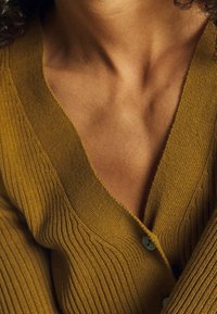 Senapsgul rippad cardigan med en djup V-hals, utrustad med två knappar och en mjuk textur. Tyget verkar lätt och gemütligt.