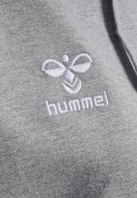 Tissu de sweat-shirt gris avec un logo brodé blanc représentant un design de abeille stylisé et le nom de la marque "hummel" en dessous.