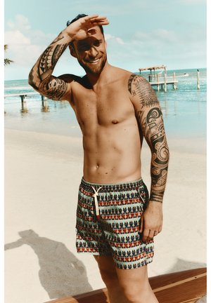 Homme torse nu avec des tatouages tribaux à manches longues protège ses yeux du soleil sur une plage de sable près de quais en bois et d'eau calme.