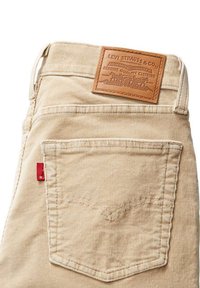 Pantalons en velours côtelé beige avec une étiquette en cuir tan à la taille et un onglet rouge sur la poche arrière. Coutures distinctives sur la poche.