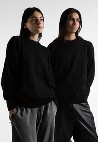 Μαύρα oversized πουλόβερ με ραμμένο λαιμόκοψη, φορεμένα πάνω από γκρι και μαύρα παντελόνια, με χαλαρές γραμμές και μίνιμαλ σχέδιο.