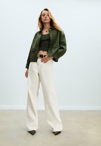 Giacca in suede verde con due tasche frontali, abbinata a un top corto verde scuro e pantaloni a palazzo crema. Presenta una cintura fantasia e scarpe con punta.