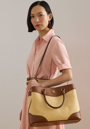 Femeie într-o rochie roz ținând o geantă tote bej cu margini din piele maro și un logo auriu RLL pe bretea.