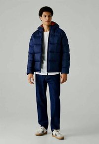Casaco de abrigo azul-marinho com capuz e design acolchoado, apresentando forro interior vermelho, combinado com jeans escuros e sapatilhas brancas.