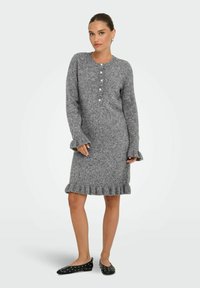 Femme portant une robe pull grise avec des détails de boutons et des poignets et un ourlet à volants, associée à des ballerines noires à clous.