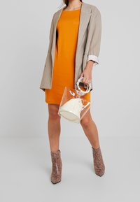 Orange dress with a relaxed fit, paired with a beige pinstriped blazer. Clear bag with a white pouch and snake print ankle boots.