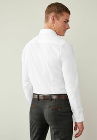 Cămașă albă cu guler standard, asortată cu pantaloni în carouri închise la culoare, având un curea maro și buzunare din spate cu nasturi.