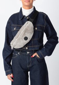 Grå crossbody-taske i kunstig pels med lynlås, der har et rundt sort logo-patch. Stropperne er sorte med justerbar hardware.